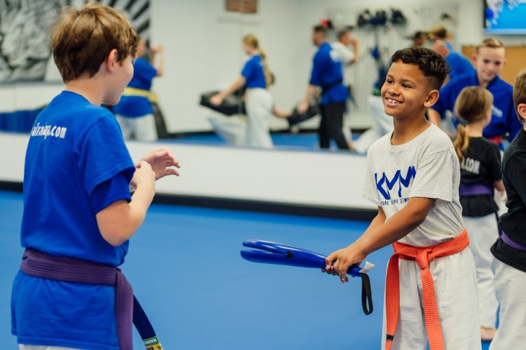 Two children in martial arts uniforms are practicing with a blue training paddle.