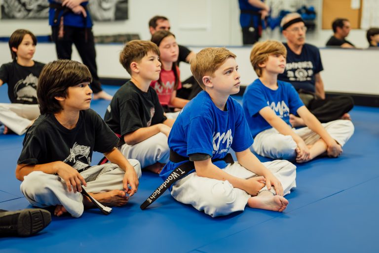 A group of children in martial arts uniforms sitting on a blue mat, listening attentively during a class