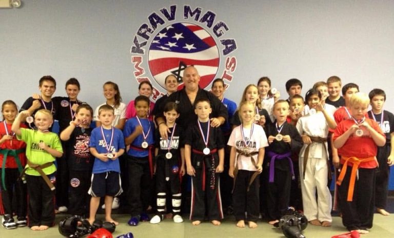 A large group of children and instructors in martial arts uniforms standing in front of a Krav Maga logo, many wearing medals