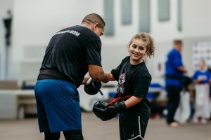 A father secures a boxing glove on his daughter as they prepare for a blackbelt test. The daughter is looking towards her side and smiling.