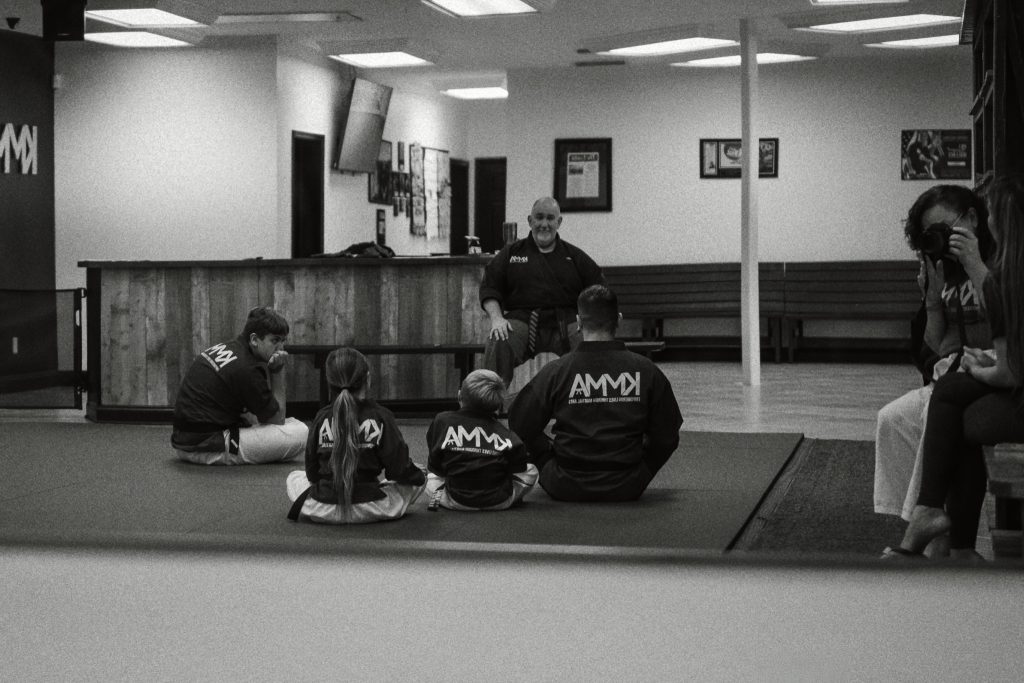 Black and white image of several students and a seated martial arts instructor in a dojo, with a person taking photos on the right side.