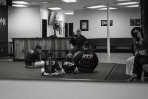 Black and white image of several students and a seated martial arts instructor in a dojo, with a person taking photos on the right side.