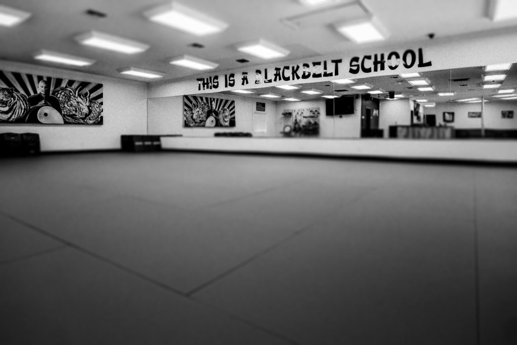 Black and white interior shot of the Krav Maga Martial Arts training floor, featuring a wide expanse of empty mats, a large mirror reflecting the ceiling lights, and the words "THIS IS A BLACKBELT SCHOOL" hanging across the top of the mirror.