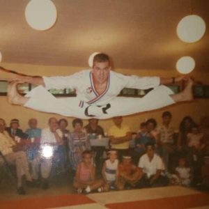 A martial artist in a white uniform with a red, white, and blue collar performs a mid-air split, holding onto his feet with his outstretched arms. Below him, an audience of seated onlookers watches the demonstration in an indoor space.