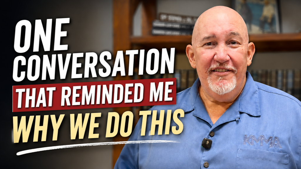 Subject: Grandmaster Stephen J. Del Castillo smiling while wearing a blue collared shirt with the KMMA logo embroidered on the chest pocket.Text Overlay: Bold text on the left side reads "ONE CONVERSATION THAT REMINDED ME WHY WE DO THIS" in white, red, and yellow lettering.Background: An office or studio setting with a wooden bookshelf displaying books and a framed picture.