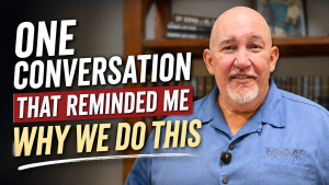 Subject: Grandmaster Stephen J. Del Castillo smiling while wearing a blue collared shirt with the KMMA logo embroidered on the chest pocket. Text Overlay: Bold text on the left side reads "ONE CONVERSATION THAT REMINDED ME WHY WE DO THIS" in white, red, and yellow lettering. Background: An office or studio setting with a wooden bookshelf displaying books and a framed picture.