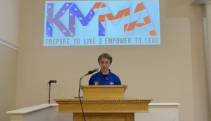 A young person wearing a blue martial arts uniform stands at a wooden podium, speaking into a microphone. Above them, a projector screen displays the text "KMMA PREPARE TO LIVE EMPOWER TO LEAD" with a red, white, and blue graphic.