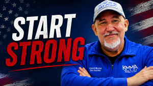 An older man with a trimmed white goatee and a friendly smile stands with his arms crossed against a dark blue background featuring weathered American flag graphics in the upper corners. He is wearing a blue zip-up jacket with "Grand Master Del Castillo" on the left chest and the KMMA logo with "PREPARE TO LIVE, EMPOWER TO LEAD" on the right. He also wears a gray baseball cap with a patch that reads "THE FUTURE LOOKS BRIGHT!". Bold white and red text on the left side of the image reads "START STRONG".