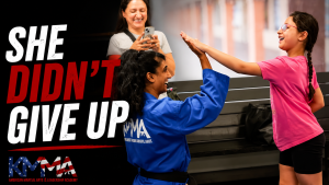 A martial arts instructor in a blue uniform and black belt gives a high-five to a young girl in a pink t-shirt and glasses, while an adult in the background smiles and takes a photo with a smartphone. Large text on the left reads "SHE DIDN'T GIVE UP" alongside the American Martial Arts Leadership Academy (KMMA) logo.