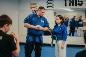 A martial arts instructor with a black belt coaches a focused youth student wearing a green belt and blue uniform. Other students are visible in the foreground and reflected in the background wall mirror.