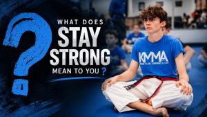 teenage martial arts student sits cross-legged on a blue mat inside a dojo, wearing a white gi, a red belt with black stripes, and a blue KMMA t-shirt that reads "Prepare to Live & Empower to Lead." Large bold text on the left asks, "What does STAY STRONG mean to you?" with other students visible in the blurred background.