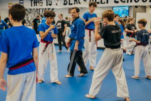 Inside a martial arts dojo with blue mats, a group of students in white uniforms and blue or black shirts practice stances. An instructor in a blue top and black pants stands in the center observing, while other students train in the background.
