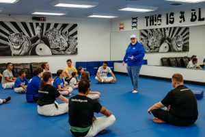Grandmaster Stephen Del Castillo stands on a blue mat inside a martial arts dojo, speaking to a diverse group of children and adults sitting cross-legged on the floor in front of him. He is wearing a blue zip-up jacket, jeans, and a baseball cap. The dojo walls feature large wall murals of a tiger and dragon flanking a portrait, a mirrored wall, and overhead white lighting.