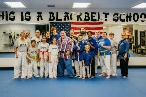 A large group of martial arts students and instructors pose together on blue mats in front of an American flag and a sign reading "This is a black belt school".