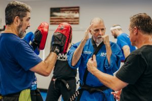 Inside a martial arts training studio, a senior instructor in a blue uniform and black belt stands defensively with raised hands while another practitioner in a black t-shirt guides him. To the left, a student in a blue t-shirt and black belt holds up red Century focus pads for partner training. In the background, other students train under an American flag hanging on the wall.