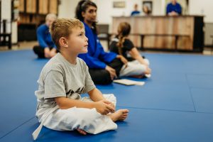 A young boy with short blonde hair and a gray t-shirt sits cross-legged on a blue martial arts mat, looking attentively forward with a white belt tied around his waist. In the blurred background, other students in martial arts uniforms are seated on the mat inside a training studio.