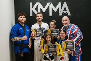 An instructor in a blue martial arts uniform with a black belt stands on the left next to a family of four smiling and holding up their Yellow Belt KMMA certificates and folded yellow belts, while an instructor in a patriotic red, white, and blue uniform stands on the right beneath a KMMA wall logo.