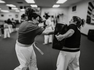 Black and white action photograph of two adult women practicing martial arts in a bright dojo. On the left, a woman wearing a t-shirt and white uniform pants throws a strong punch forward. On the right, her training partner holds a large black strike shield absorbing the impact, her expression focused as she braces for the hit. Other students and instructors are softly blurred in the background.