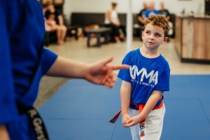 A young boy wearing a blue KMMA t-shirt, white martial arts pants, and an orange belt stands on blue mats, attentively listening as an instructor points out-of-frame to his left.