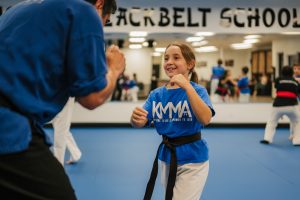 A young female martial arts student wearing a blue KMMA t-shirt, white uniform pants, and a black belt practices a martial arts stance while facing an instructor inside a dojo with blue mats and mirrored walls.