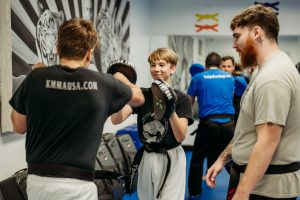 Two young martial arts students training inside a dojo, with one holding focus mitts and smiling while blocking a strike, and an instructor with a reddish beard observing closely nearby.