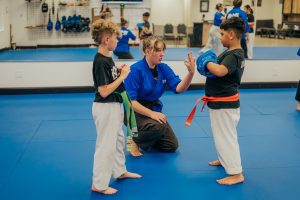 In a bright martial arts dojo with blue floor mats and mirrored walls, a teenage or young adult instructor wearing a blue uniform top, black pants, and a black belt is kneeling on the floor. She is gesturing with her hand while talking to two children standing across from her. On the left, a young boy with curly blonde hair wears a black t-shirt, white uniform pants, and a green belt. On the right, another young boy with dark hair wears a black t-shirt, white pants, an orange belt, and blue hand pads. Other students and instructors are visible in the background out of focus.