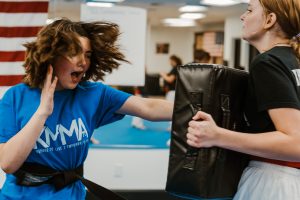 A young martial artist in a blue KMMA t-shirt and black belt throws a powerful palm strike into a black target shield held by a training partner, with her hair flying mid-motion as she shouts.