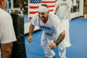 A male martial artist with a focused expression strains intensely while pulling a resistance band during training inside a dojo, wearing a white KMMA t-shirt, white belt, and a headband, with an American flag displayed in the background.