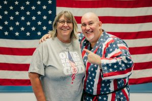 Grandmaster Stephen Del Castillo stands smiling next to his wife Barbara who is wearing a grey t-shirt that reads "ONE NATION UNDER GOD," with both posing in front of a large American flag.