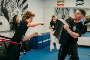 A student in a black uniform practices a resistance punch while pulling against a red band, throwing a strike toward an instructor holding a heavy training shield.