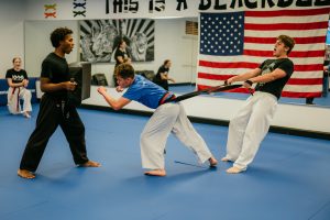 A martial arts student performs a resistance punch, straining forward while another student holds back a resistance band wrapped around their waist, as a third student holds a training pad nearby.
