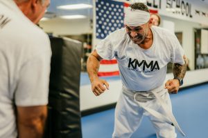 A martial arts student wearing a white KMMA t-shirt, white headband, and white belt strains intensely with clenched fists during a training drill in front of an American flag.