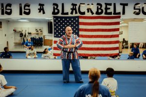 Grandmaster Stephen Del Castillo stands on a blue mat inside a martial arts school, wearing a patterned uniform and addressing seated students in front of a large American flag hung on a mirrored wall.