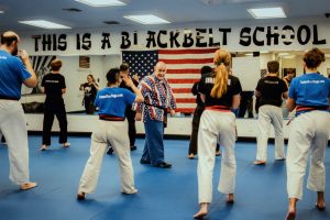 Students in uniforms practice techniques on a blue mat inside a dojo while Grandmaster Stephen Del Castillo walks among them, beneath a large American flag and the painted words "THIS IS A BLACK BELT SCHOOL".