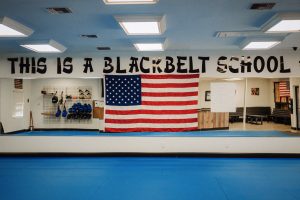 n interior view of a martial arts dojo with blue training mats on the floor, featuring a large American flag hung on a mirrored wall below bold black lettering painted near the ceiling that reads "THIS IS A BLACK BELT SCHOOL".