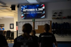 tudents face a wall-mounted TV screen displaying a patriotic memorial graphic that reads "SEPTEMBER 11, 2001 WE WILL NEVER FORGET."