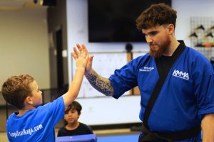 hihan Winston in a blue KMMA uniform giving a high-five to a young boy wearing a matching blue KMMA t-shirt inside the martial arts dojo.