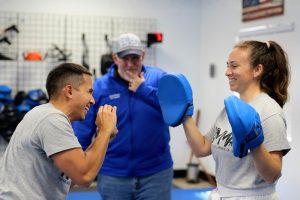 A smiling woman in a gray KMMA t-shirt and white belt wears blue focus mitts while practicing martial arts, laughing alongside a male partner in a matching gray shirt, as Grandmaster Del Castillo in a blue jacket and cap looks on from the background.