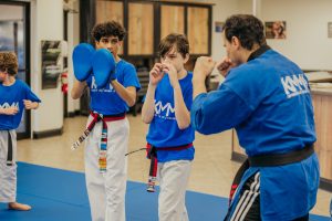 Inside a martial arts dojo, a young male student in a blue t-shirt and white pants with a black belt practices a defensive guard stance. He faces an instructor wearing a blue martial arts top who holds up his fists in a sparring posture. To the left, another student holds blue kick shields, while a younger child practices in the background on blue mats.