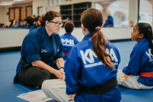 An instructor in a blue martial arts uniform sits cross-legged on a dojo mat, speaking with two younger students who are also seated in uniforms facing her.