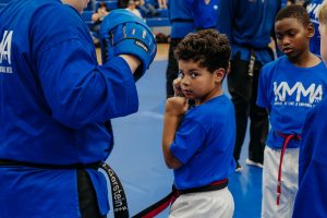 A young martial arts student with curly brown hair wearing a blue t-shirt, white pants, and a black belt with red trim practicing a martial arts stance on a blue mat, looking focused toward the camera while an instructor holds a blue hand pad nearby