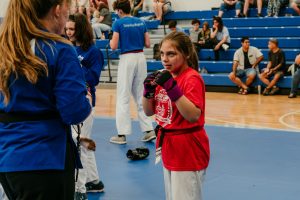 A young martial arts student wearing a red graphic T-shirt, white pants, a red belt, and padded sparring gloves stands in a focused fighting stance on a blue gym mat. In the foreground, an instructor in a blue uniform faces her, while other students and spectators seated on blue bleachers fill the background.