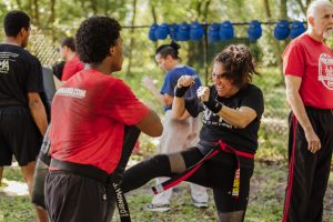 A woman with a determined expression delivers a side kick toward a training partner holding a pad during an outdoor martial arts class, while other participants and instructors train nearby against a backdrop of trees.