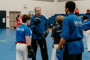 An instructor wearing a blue uniform and black belt with focus mitts on his hands talks to several students in a gymnasium with blue mats and bleachers in the background.