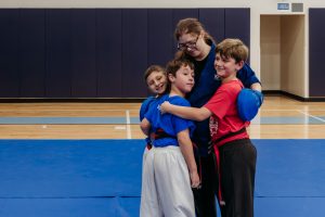 An instructor wearing glasses and a blue martial arts uniform warmly hugs and embraces three young students in a gymnasium with blue training mats and a wooden floor.   