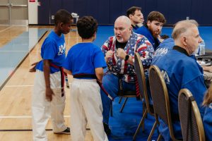 In a gymnasium with wooden floors and blue mats, two young martial artists wearing blue t-shirts, white pants, and black belts stand facing Grandmaster Del Castillo, who is seated. The Grandmaster, who is wearing a distinctive American flag patterned martial arts top, gestures animatedly while talking to the children. Several other instructors and students in blue uniforms sit at tables and chairs along the right side of the frame, watching the interaction.