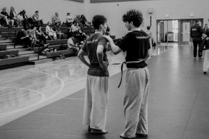 A black-and-white photo showing two young martial students in t-shirts and loose pants standing on a mat during an event. The student on the right has his hand on the other student's shoulder in a gesture of support. Both wear shirts printed with "TampaKravMaga.com". Spectators sit on bleachers in the gymnasium background
