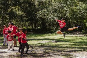 Outdoor photo of a young male martial artist captured in mid-air performing an impressive flying kick. He is wearing a red "Krav Camp" t-shirt and black shorts, suspended high above a grassy area backed by dense green foliage. To the left, a small group of martial arts students and an instructor in matching red shirts watch his demonstration.