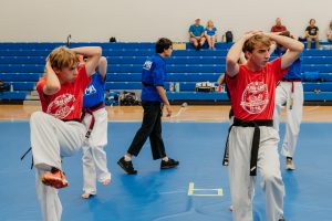 wo young martial artists in red Krav Camp shirts and black belts perform a high knee strike on a blue mat inside a gymnasium, with instructors and spectators visible in the background.