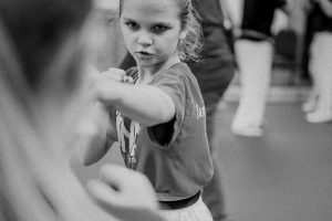 Black-and-white, close-up action photograph of a young martial arts student intensely focused as she executes a defensive block or strike in class, with another student blurred in the foreground and instructors visible in the background.